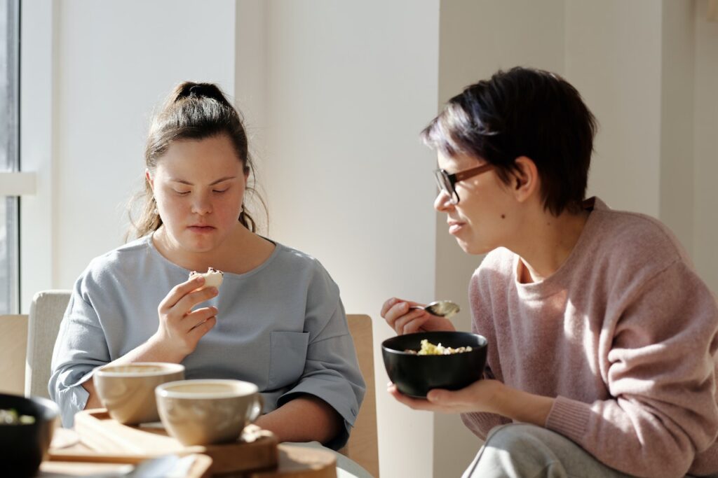 A woman looking after and eating with another woman with Down Syndrome.
